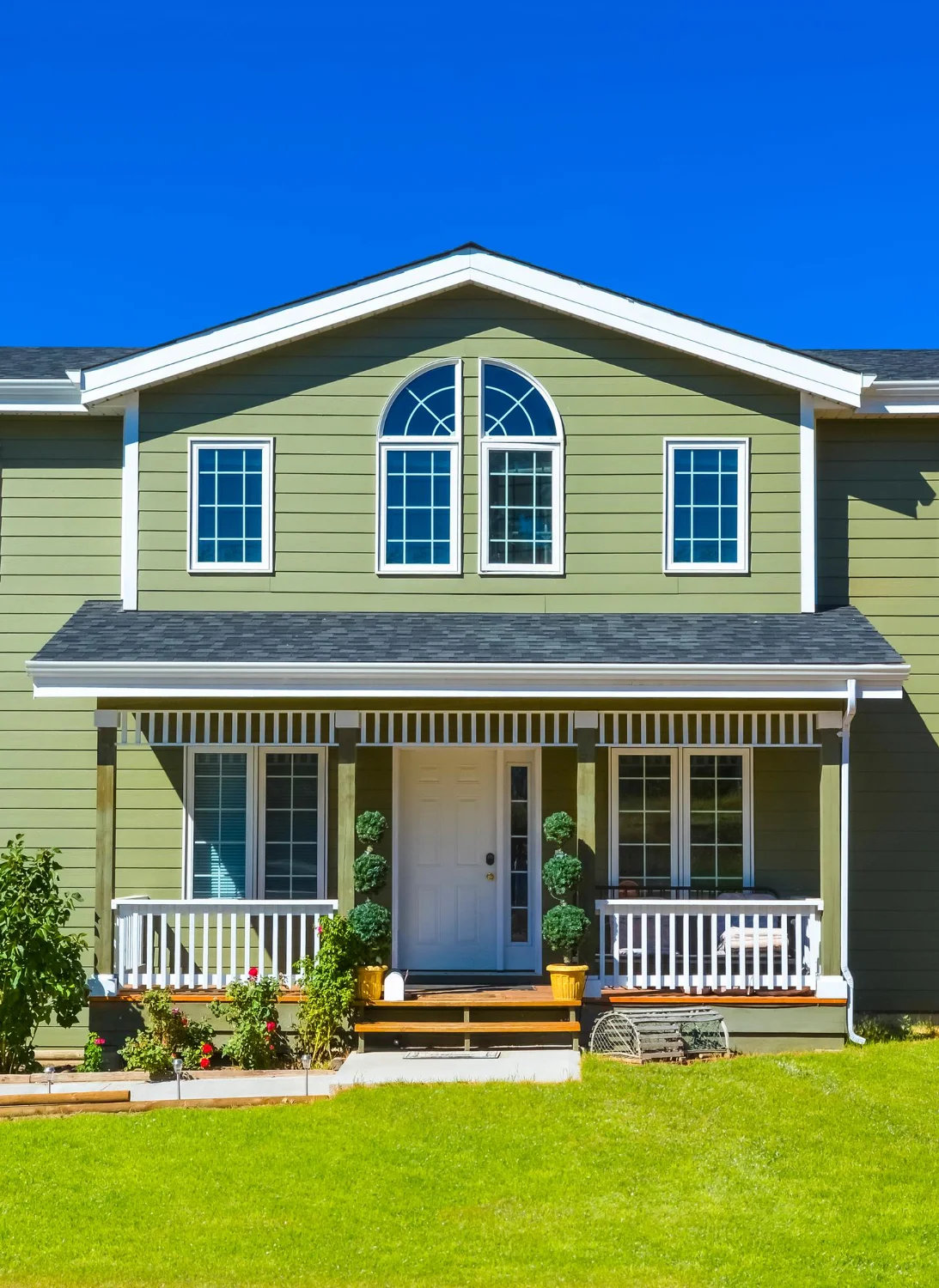 A large green house featuring a charming white porch, surrounded by a lush garden